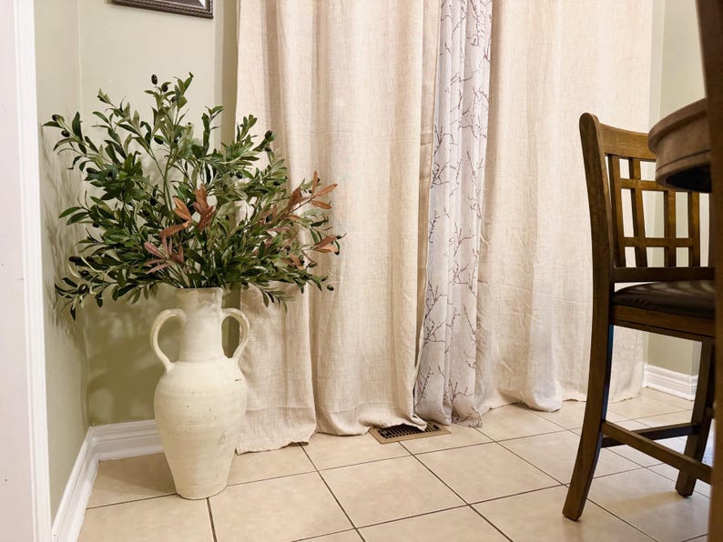 May include: A white ceramic vase with two handles, filled with a green and brown olive branch arrangement, sits in front of a beige linen curtain with a sheer white curtain behind it. The vase is on a tiled floor.
