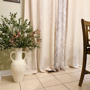 May include: A white ceramic vase with two handles, filled with a green and brown olive branch arrangement, sits in front of a beige linen curtain with a sheer white curtain behind it. The vase is on a tiled floor.
