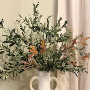 May include: A bouquet of olive branches with green leaves and small black olives in a white ceramic vase with two handles.