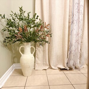 May include: A white ceramic vase with two handles, filled with a green and brown leafy plant. The vase is standing on a light brown tiled floor in front of a white curtain with a floral pattern.
