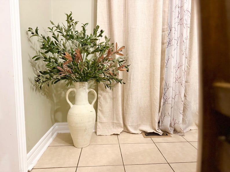 May include: A white ceramic vase with two handles, filled with a green and brown leafy plant. The vase is standing on a light brown tiled floor in front of a white curtain with a floral pattern.