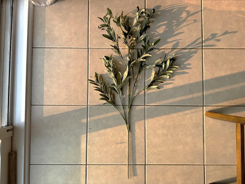 May include: A single artificial olive branch with green leaves and small black olives. The branch is lying flat on a light brown tiled floor.