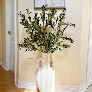 May include: A white ceramic vase with two handles, filled with a large arrangement of green olive branches with small black olives.
