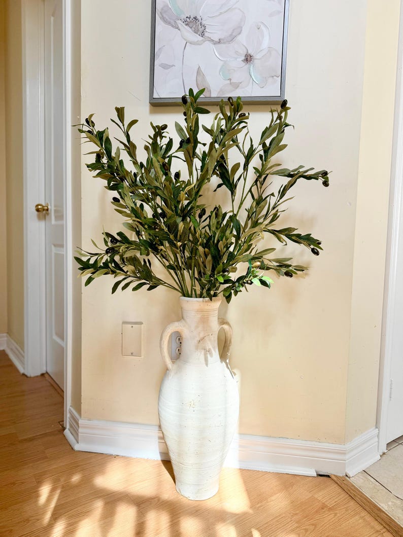 May include: A white ceramic vase with two handles, filled with a large arrangement of green olive branches with small black olives.