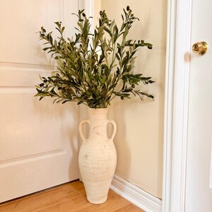 May include: A white ceramic vase with two handles, filled with a green olive branch with small black olives.
