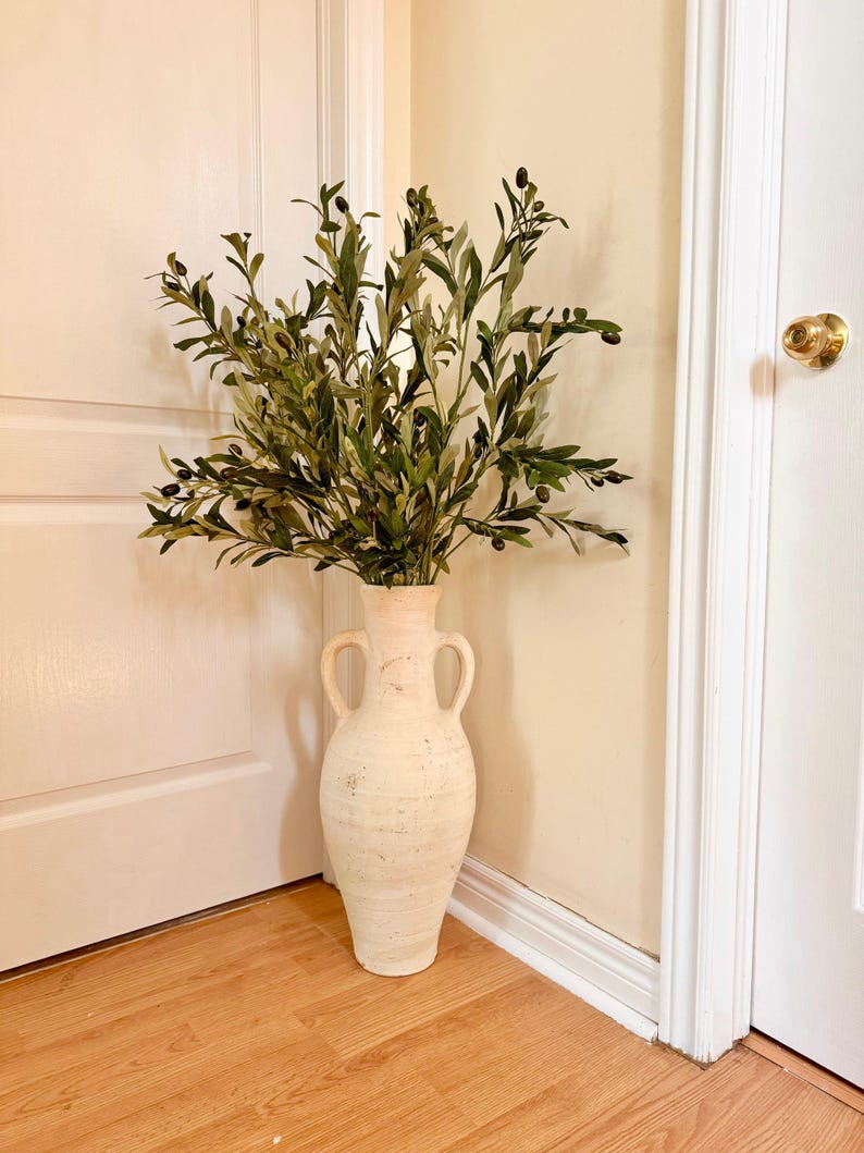 May include: A white ceramic vase with two handles, filled with a green olive branch with small black olives.