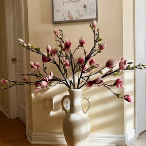 May include: A white ceramic vase with two handles, filled with pink magnolia flower stems. The vase is sitting on a wooden floor in front of a white wall.