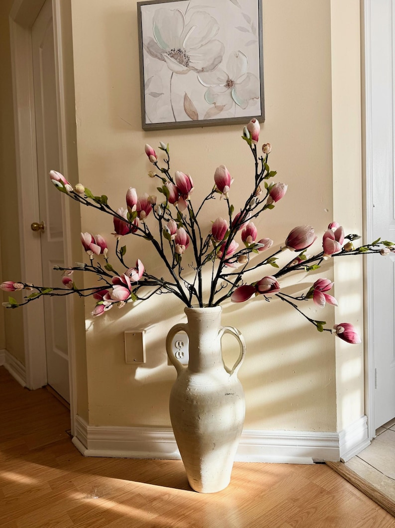 May include: A white ceramic vase with two handles, filled with pink magnolia flower stems. The vase is sitting on a wooden floor in front of a white wall.