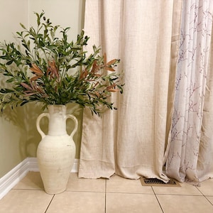 May include: A white ceramic vase with two handles, filled with a green leafy plant with small brown berries. The vase is sitting on a tiled floor in front of a beige curtain.