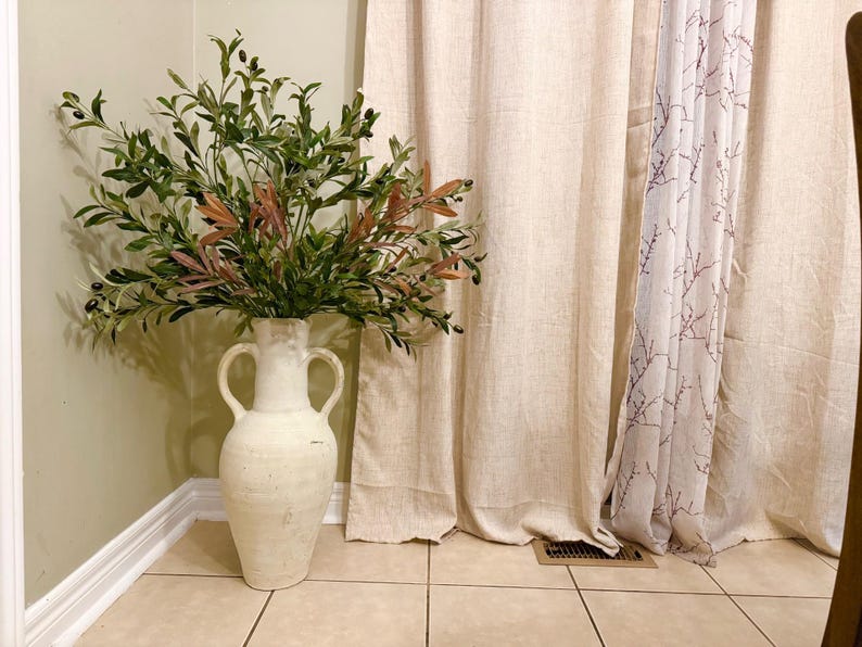 May include: A white ceramic vase with two handles, filled with a green leafy plant with small brown berries. The vase is sitting on a tiled floor in front of a beige curtain.