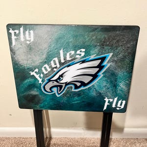 May include: A folding table with a teal and white marbled design. The Philadelphia Eagles logo is centered on the table, with the words "Fly Eagles Fly" in white script. The table has black metal legs.