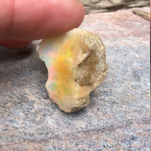 Puede incluir: Una piedra preciosa de &oacute;palo natural en bruto con una base blanca lechosa y destellos iridiscentes de amarillo, naranja y verde. El &oacute;palo es una pieza en bruto sin pulir con una forma irregular y rugosa.