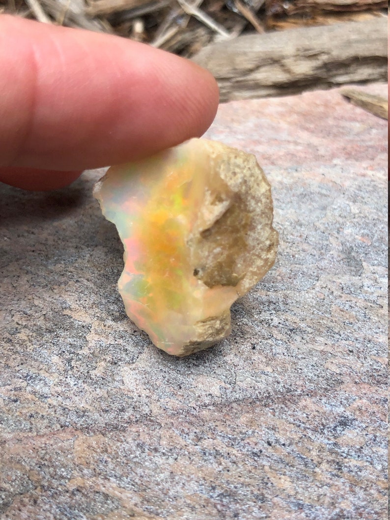 Puede incluir: Una piedra preciosa de &oacute;palo natural en bruto con una base blanca lechosa y destellos iridiscentes de amarillo, naranja y verde. El &oacute;palo es una pieza en bruto sin pulir con una forma irregular y rugosa.