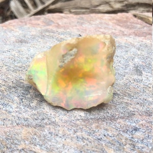 Puede incluir: Un &oacute;palo blanco lechoso rugoso con destellos iridiscentes de verde, amarillo y naranja. El &oacute;palo est&aacute; sentado sobre una roca gris rugosa.