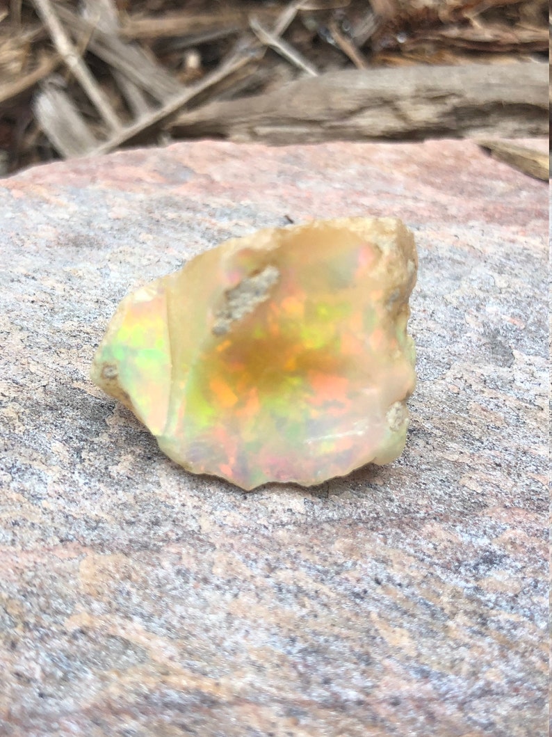 Puede incluir: Un &oacute;palo blanco lechoso rugoso con destellos iridiscentes de verde, amarillo y naranja. El &oacute;palo est&aacute; sentado sobre una roca gris rugosa.