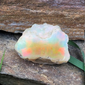 Puede incluir: Una piedra preciosa de &oacute;palo en bruto, blanca, con colores arco&iacute;ris iridiscentes, sentada sobre una roca marr&oacute;n.