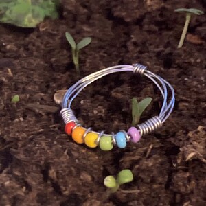 May include: A handmade wire ring with a rainbow of colorful beads. The ring features red, orange, yellow, green, blue, and purple beads. The ring is set against a backdrop of dark soil with small green plants.