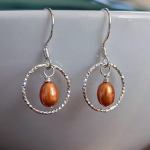 May include: A pair of silver dangle earrings. Each earring features a textured silver hoop with a small, oval, orange-brown pearl suspended in the center. The earrings are displayed against a blurred white background.