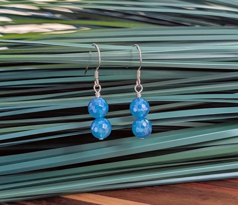 May include: A pair of drop earrings featuring two faceted, round, bright blue beads on each earring. The beads are suspended from silver-toned metal fishhook wires, creating a simple and symmetrical design with a reflective, textured surface.