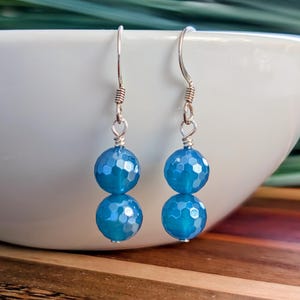 May include: A pair of drop earrings featuring two faceted, round, bright blue beads stacked vertically on each piece. The beads are suspended from silver-toned metal ear wires with a simple hook design and a small coiled detail at the top.