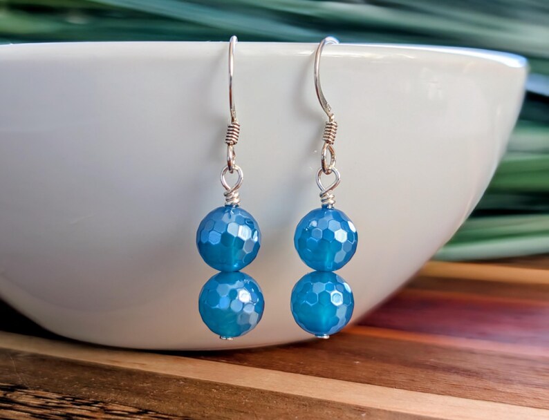 May include: A pair of drop earrings featuring two faceted, round, bright blue beads stacked vertically on each piece. The beads are suspended from silver-toned metal ear wires with a simple hook design and a small coiled detail at the top.