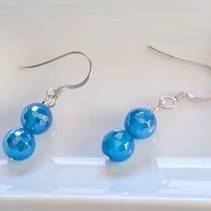 May include: A pair of drop earrings featuring two faceted, bright blue spherical beads suspended from silver-toned metal fishhook wires. Each earring consists of a smaller bead positioned above a larger bead, connected by silver-toned metal rings.