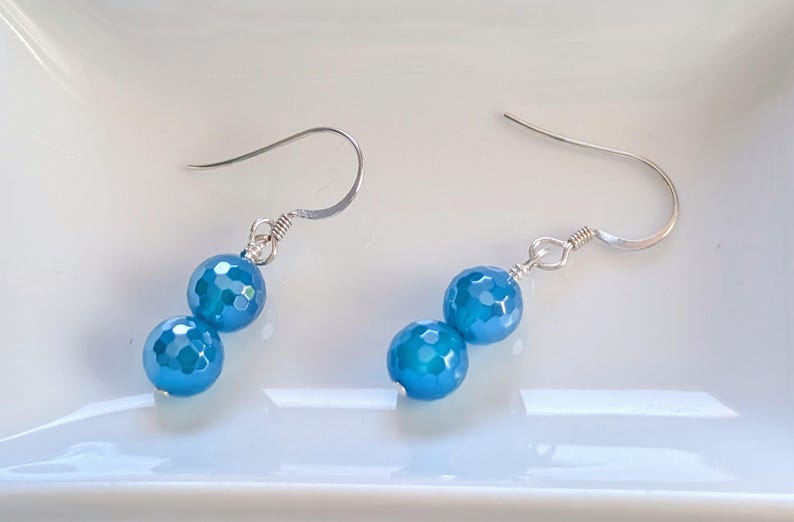 May include: A pair of drop earrings featuring two faceted, bright blue spherical beads suspended from silver-toned metal fishhook wires. Each earring consists of a smaller bead positioned above a larger bead, connected by silver-toned metal rings.