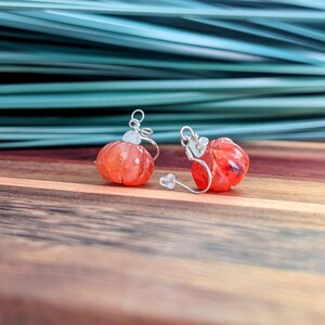 以下が含まれることがあります： シルバーカラーの金属製フックと透明なプラスチック製キャッチが付いた、オレンジ色のカボチャ型イヤリングのペア。イヤリングは、小さなカボチャの外観を模したリブ付きの丸みを帯びたデザインで、光沢のある仕上げが施されています。