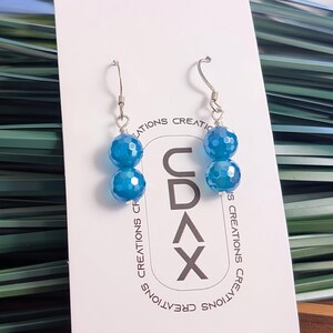 May include: A pair of dangle earrings featuring two faceted, bright blue spherical beads each, suspended from silver-toned metal hooks. The earrings are displayed on a white rectangular card printed with the text CDAX CREATIONS.