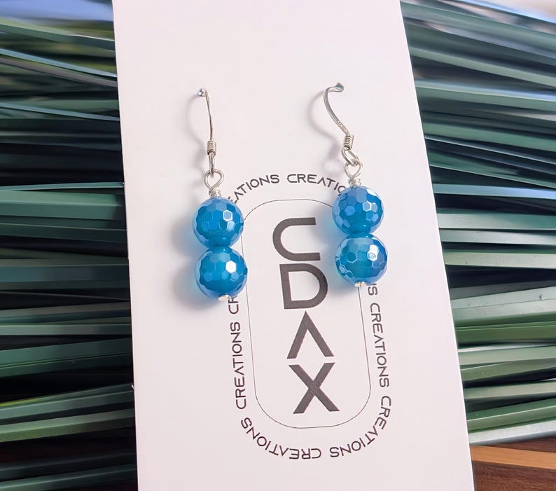 May include: A pair of dangle earrings featuring two faceted, bright blue spherical beads each, suspended from silver-toned metal hooks. The earrings are displayed on a white rectangular card printed with the text CDAX CREATIONS.