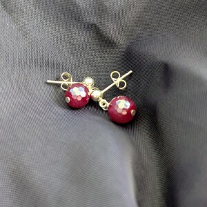 K&ouml;nnte beinhalten: Ein Paar Ohrstecker mit facettierten, tiefroten, kugelf&ouml;rmigen Perlen an kleinen goldfarbenen Metallstiften. Jeder Ohrring besteht aus einer runden, texturierten roten Perle, die an einer glatten goldfarbenen Kugel h&auml;ngt und mit einem Verschluss gesichert ist.