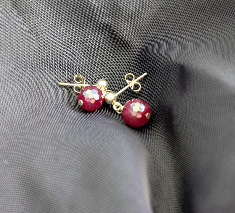 K&ouml;nnte beinhalten: Ein Paar Ohrstecker mit facettierten, tiefroten, kugelf&ouml;rmigen Perlen an kleinen goldfarbenen Metallstiften. Jeder Ohrring besteht aus einer runden, texturierten roten Perle, die an einer glatten goldfarbenen Kugel h&auml;ngt und mit einem Verschluss gesichert ist.