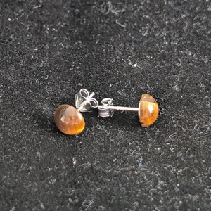 May include: A pair of stud earrings featuring round, polished tiger's eye gemstones in a warm golden-brown hue. The stones are set in silver-toned metal posts with matching butterfly-style earring backs, showcasing a smooth, domed surface.
