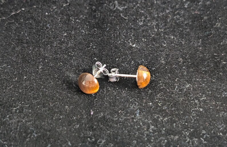 May include: A pair of stud earrings featuring round, polished tiger's eye gemstones in a warm golden-brown hue. The stones are set in silver-toned metal posts with matching butterfly-style earring backs, showcasing a smooth, domed surface.