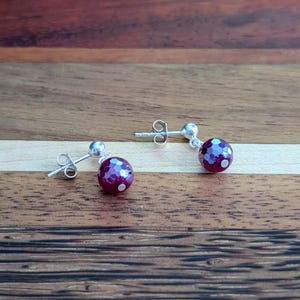 K&ouml;nnte beinhalten: Ein Paar Ohrstecker mit runden, facettierten, tief magentafarbenen Perlen, die eine schimmernde, vielschichtige Oberfl&auml;che aufweisen. Jede Perle ist an einem silberfarbenen Metallstift mit einem kleinen silbernen Kugelakzent befestigt.