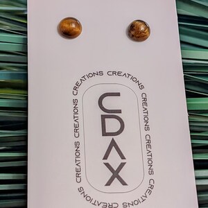 May include: A pair of round, brown tiger's eye stud earrings mounted on a white display card. The card features the text CDAX vertically in a rounded rectangle, surrounded by the repeated word CREATIONS in a circular pattern around the logo.
