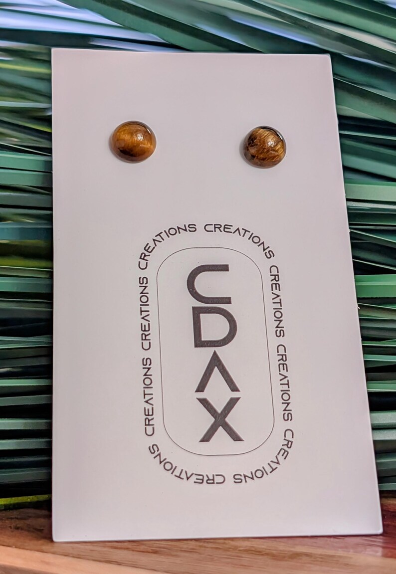 May include: A pair of round, brown tiger's eye stud earrings mounted on a white display card. The card features the text CDAX vertically in a rounded rectangle, surrounded by the repeated word CREATIONS in a circular pattern around the logo.