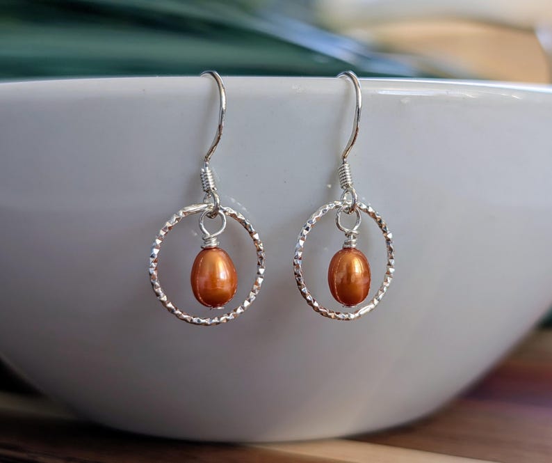 May include: A pair of silver dangle earrings. Each earring features a textured silver hoop with a small, oval, orange-brown pearl suspended in the center. The earrings are displayed against a blurred white background.