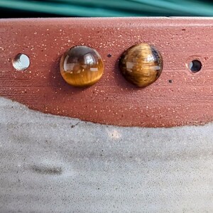 May include: Two round, polished tiger's eye gemstones with a chatoyant golden-brown luster are set against a textured, two-toned ceramic surface. The upper portion of the ceramic is a speckled reddish-brown, while the lower section is a light gray.