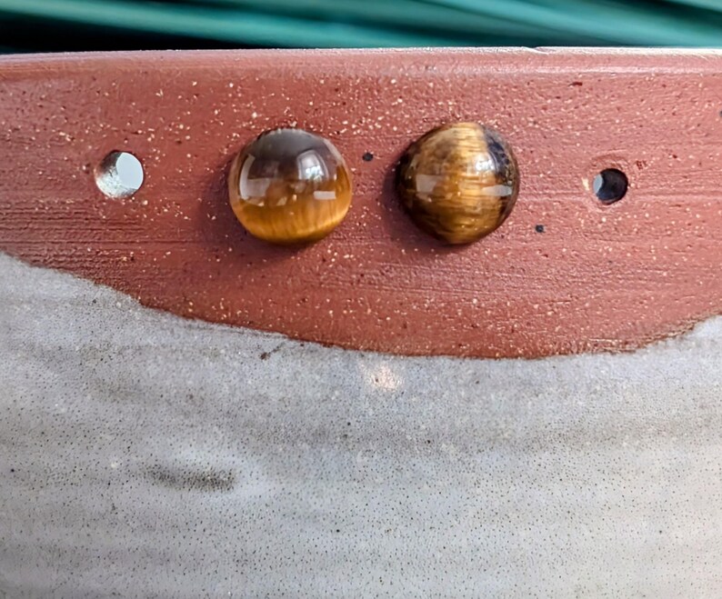May include: Two round, polished tiger's eye gemstones with a chatoyant golden-brown luster are set against a textured, two-toned ceramic surface. The upper portion of the ceramic is a speckled reddish-brown, while the lower section is a light gray.