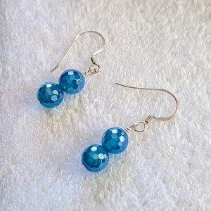 May include: A pair of drop earrings featuring two faceted, round, bright blue beads suspended from silver-toned metal hooks. Each earring consists of a smaller bead positioned above a slightly larger bead, connected by silver-toned wire components.