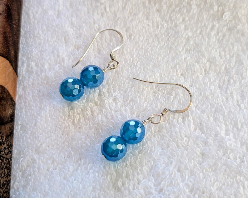 May include: A pair of drop earrings featuring two faceted, round, bright blue beads suspended from silver-toned metal hooks. Each earring consists of a smaller bead positioned above a slightly larger bead, connected by silver-toned wire components.