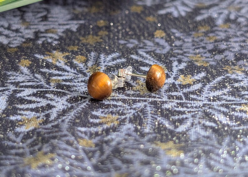 May include: A pair of stud earrings featuring round, polished tiger's eye gemstones with a chatoyant golden-brown luster. The stones are set on silver-toned metal posts with matching butterfly clutch backings, displaying a smooth, domed surface.