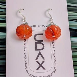 以下が含まれることがあります： CDAX CREATIONSの文字が印刷された白い台紙に取り付けられた、オレンジ色のカボチャ型イヤリング。銀色のフックが付いており、カボチャのビーズにはリブ状の質感と上部に小さな緑色の葉のディテールが施されています。