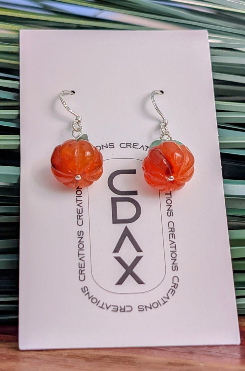 以下が含まれることがあります： CDAX CREATIONSの文字が印刷された白い台紙に取り付けられた、オレンジ色のカボチャ型イヤリング。銀色のフックが付いており、カボチャのビーズにはリブ状の質感と上部に小さな緑色の葉のディテールが施されています。
