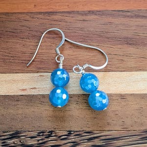 May include: A pair of dangle earrings featuring two faceted, bright blue spherical beads each, suspended from silver-toned metal ear wires. The beads have a textured, multi-faceted surface that reflects light, and they are connected by thin silver-toned wire.