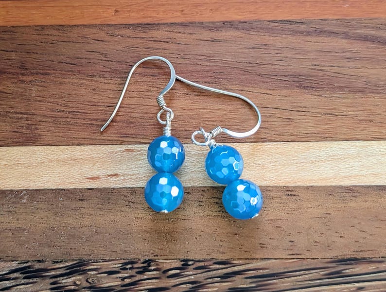 May include: A pair of dangle earrings featuring two faceted, bright blue spherical beads each, suspended from silver-toned metal ear wires. The beads have a textured, multi-faceted surface that reflects light, and they are connected by thin silver-toned wire.