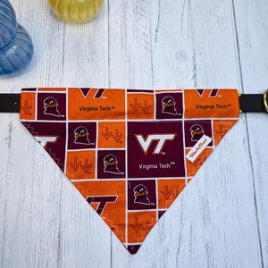 May include: Orange and maroon dog bandana with a Virginia Tech design. The bandana features the Virginia Tech logo, the words "Virginia Tech", and a cartoon duck. The bandana is on a black collar.