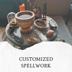 Può includere: Un tavolo rotondo con una ciotola in legno, una conchiglia riempita di erbe, vari cristalli, una cupola di vetro con un teschio e due candele accese. La superficie del tavolo mostra il testo CUSTOMIZED SPELLWORK. L'insieme include pietre nere, marroni e trasparenti.