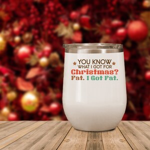May include: A white insulated wine tumbler with a silver rim. The tumbler has the text "YOU KNOW WHAT I GOT FOR Christmas? Fat. I Got Fat." in various colors. The background is a blurred Christmas tree.